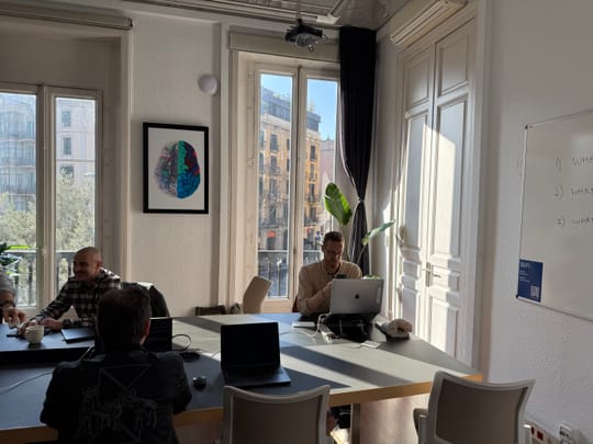 The main Eixample 46 coworking room — a long communal table with members at work, sunlight pouring through tall French windows onto the original wood floor
