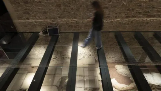 Archaeologists examining ancient Roman paving stones in an excavation site in Barcelona, with historic buildings in the backg