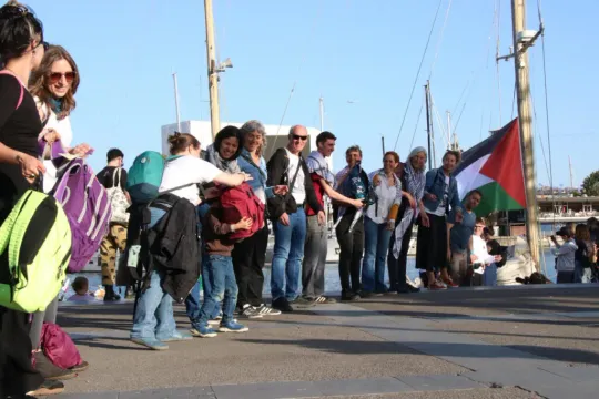 Humanitarian flotilla vessels departing from Barcelona port, with people waving from the docks.