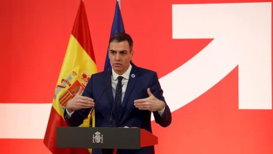 Spanish Prime Minister Pedro Sánchez speaking at a podium with a Spanish flag in the background.