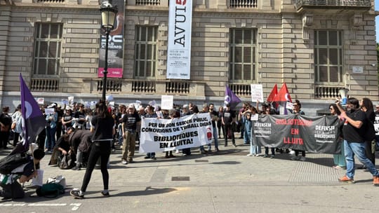Library workers marching in a protest through Barcelona, holding banners and signs, with city buildings in the background.