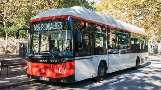 A red and white Barcelona V5 bus driving on a city street, with buildings in the background.