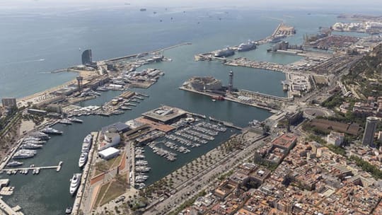 An aerial view of Barcelona's Port Vell showing new waterfront developments, a large esplanade, and boats in the marina.