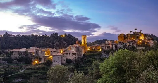 A wooden suspension bridge leading to a medieval village with stone houses and a church tower, surrounded by lush trees.