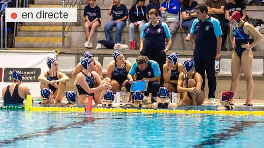 Women's water polo players in a pool, one in a white cap with 'Sant Andreu' written on it, another in a blue cap, competing f