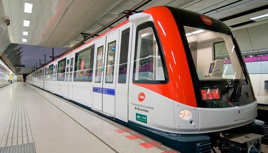 A crowded Barcelona Metro train carriage with diverse passengers standing and sitting.