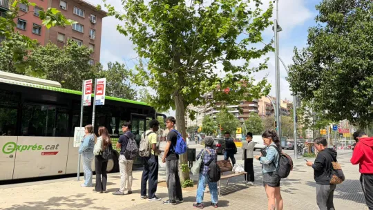 People waiting in a long queue at a busy interurban bus stop in Barcelona, with city buildings in the background.