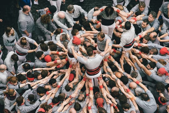 A tall human tower, or castell, being built by a group of people in a public square in Barcelona, with spectators watching.