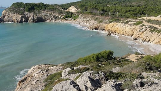 A picturesque cove with clear blue water, sandy beach, and green cliffs, with a train passing in the background.
