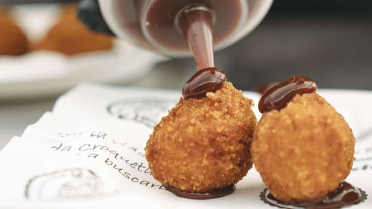 A close-up of various golden-brown croquettes on a plate, some with visible fillings, ready for tasting at a food festival.