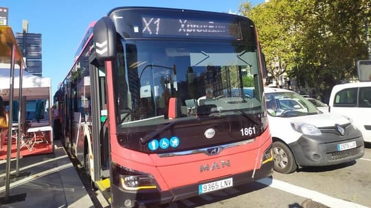A modern red and white TMB express bus driving on a street in Barcelona, with city buildings in the background.