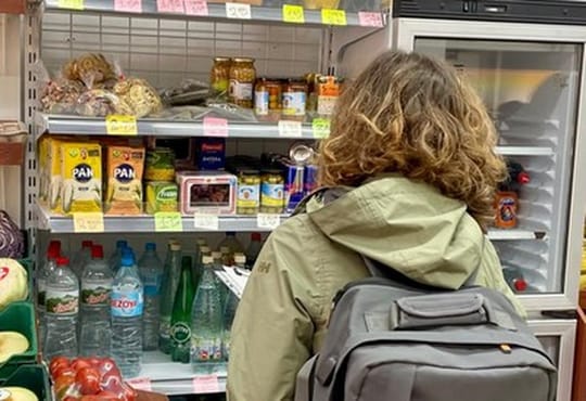 A brightly lit convenience store interior with shelves stocked with various food and household products, a cashier visible in