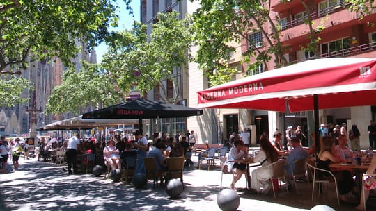 Outdoor dining terraces on Avinguda de Gaudí with the Sagrada Família in the background under a clear sky.