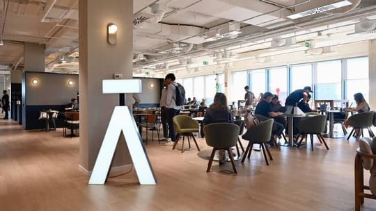 Modern, brightly lit coworking space interior with desks, chairs, and people working, showing a vibrant office environment.