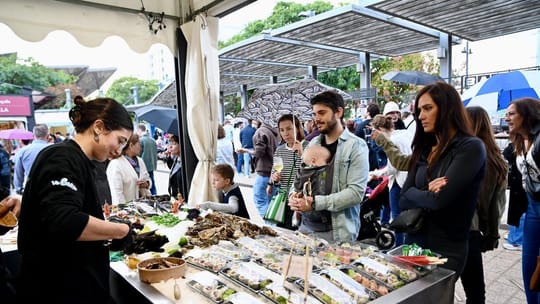 A bustling outdoor market scene at Plaça de les Glòries Catalanes, with various stalls, people browsing, and a festive atmosp