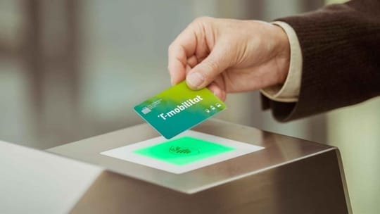 A hand holding a Barcelona Metro T-Mobilitat card, with a blurred metro station in the background.