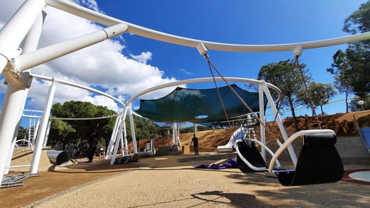 Children playing on long, wide slides at the Plaça de les Glòries playground in Barcelona, surrounded by green spaces.