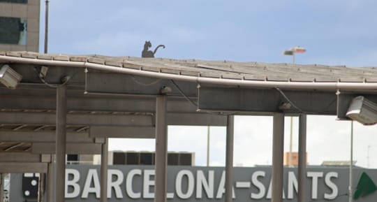 A metallic cat sculpture sitting on a concrete roof with the Sants railway station in the background, under a clear sky.