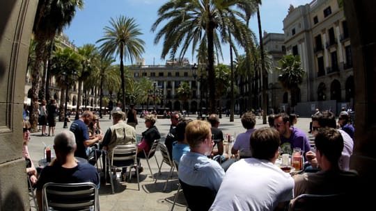 Outdoor restaurant terraces line a busy street in Barcelona, with pedestrians walking past.