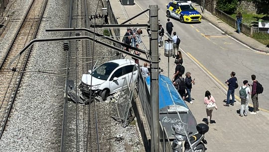 A damaged car on railway tracks next to a collapsed wall, with emergency services present.
