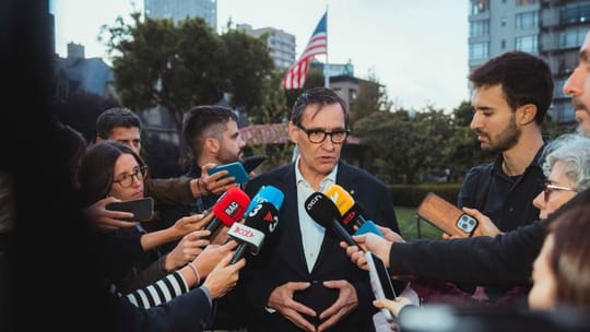 Catalan President Salvador Illa speaking at a podium with flags in the background during his visit to California.