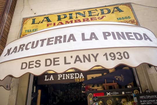 Interior of La Pineda charcuterie in Barcelona, showing cured hams, cheeses, and a customer at a marble counter.