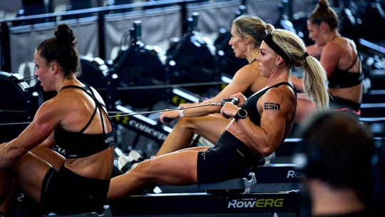 Athletes competing in a Hyrox fitness race, pushing a weighted sled across a floor, with other workout stations visible in th