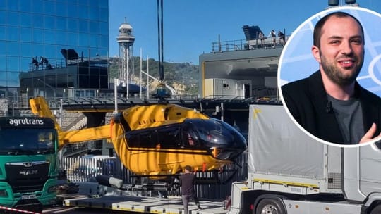 A large yellow helicopter suspended by a crane over a superyacht support vessel in Barcelona's Marina Port Vell.