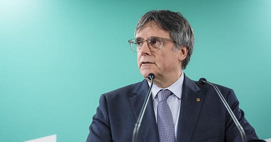 Carles Puigdemont speaking at a political rally with Junts per Catalunya flags in the background.