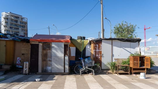 Improvised shacks made of scrap metal and wood, with laundry hanging outside, in a dusty, open field next to railway tracks i