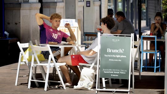 A modern cafe with outdoor seating on a tree-lined street in Poblenou, Barcelona, with people enjoying brunch.