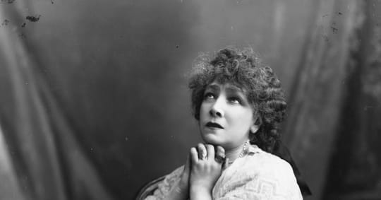 Sarah Bernhardt, an elderly woman with curly hair, sitting in an armchair, being carried by attendants at a train station pla