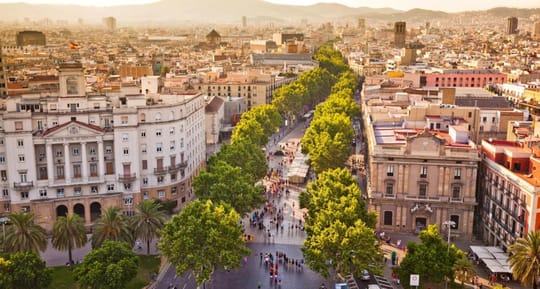 People walking along La Rambla in Barcelona, with trees and historic buildings visible, under a clear sky.