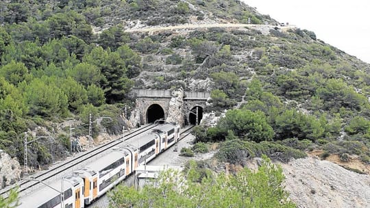 Rodalies train travelling through a tunnel near the coast, with blue sky and sea visible outside.