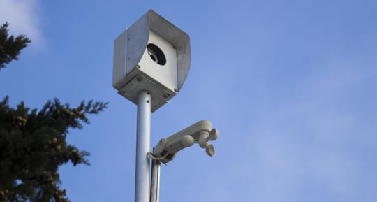 A modern CCTV camera mounted on a pole, overlooking a city street with buildings and trees.