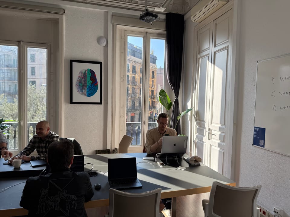 The main Eixample 46 coworking room — a long communal table with members at work, sunlight pouring through tall French windows onto the original wood floor