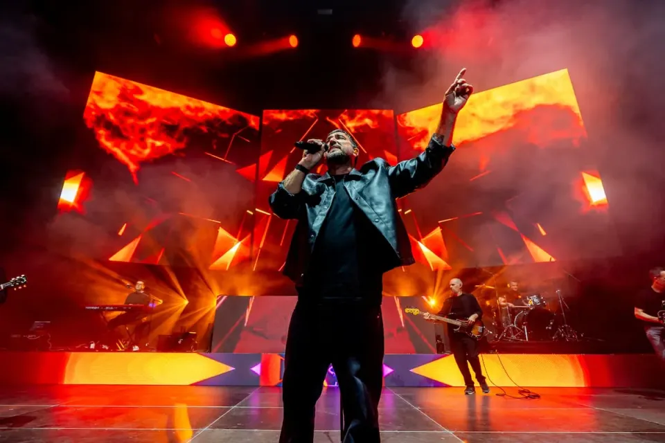 A large crowd of people watching a music concert on stage inside a brightly lit arena.
