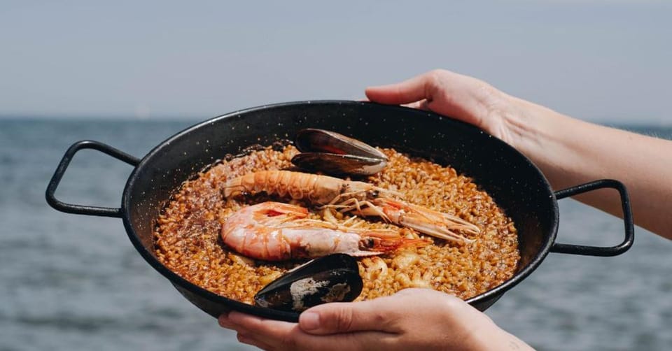 A vibrant outdoor dining terrace overlooking the Mediterranean Sea in Barcelona, with people enjoying rice dishes.