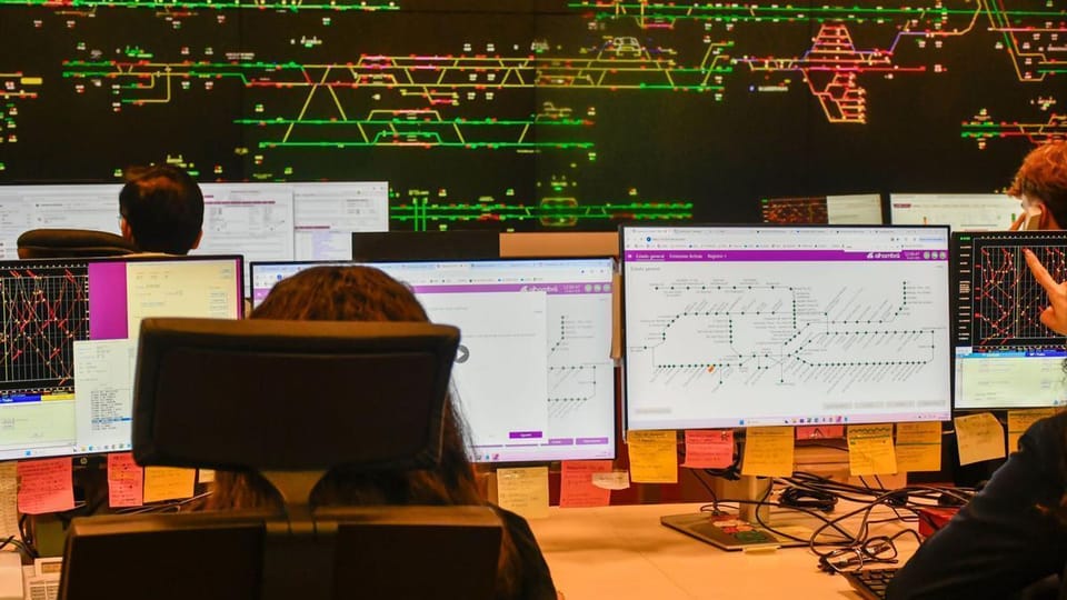 A modern train station platform with a Rodalies train arriving, showing passengers waiting and digital information screens.