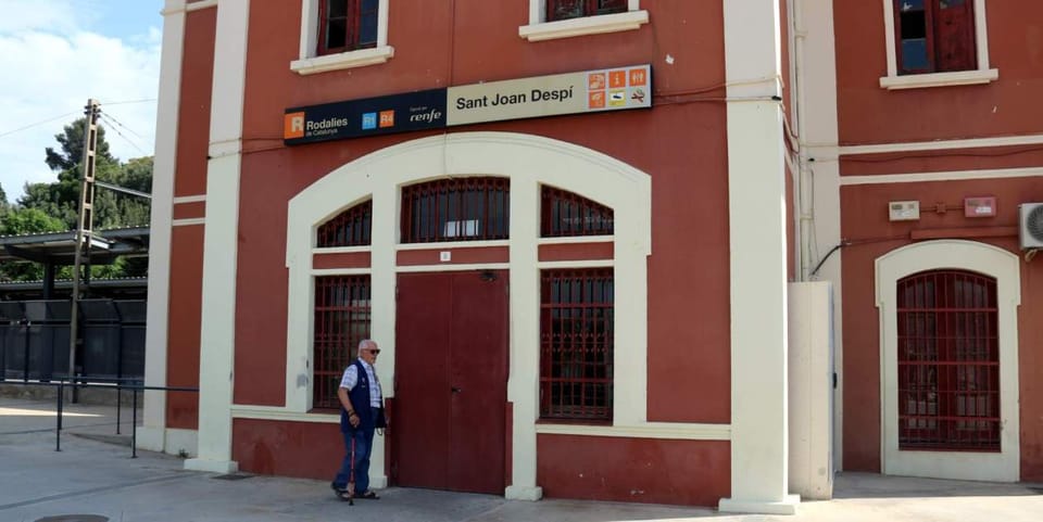 Exterior view of Sant Joan Despí train station building with a closed entrance and a Rodalies train on the tracks.