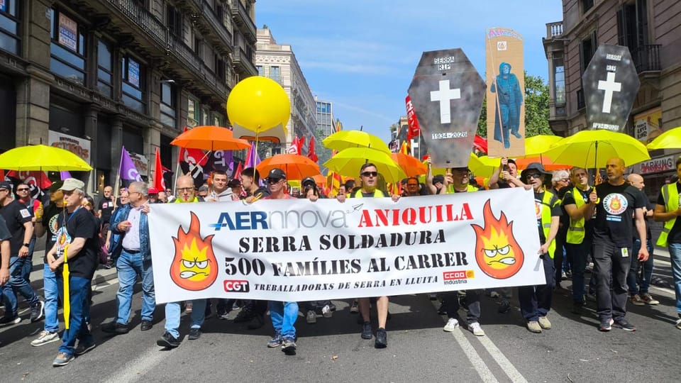 Workers protesting in a street in Barcelona, holding banners and chanting, with city buildings in the background.