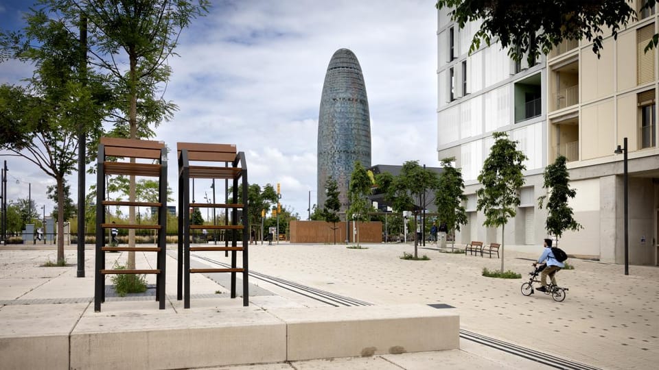 A modern sculpture in a public square in Barcelona, with historic buildings in the background under a clear sky.