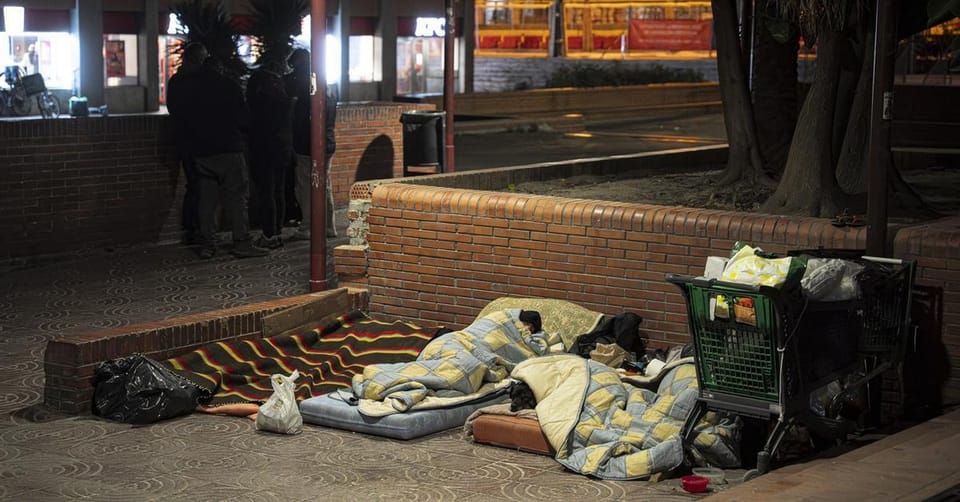 A person sitting on the street in Barcelona, with a blurred background of city life, symbolising homelessness.