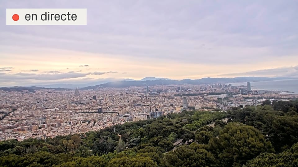 Dark clouds gather over Barcelona city, with rain starting to fall over buildings and the sea in the distance.