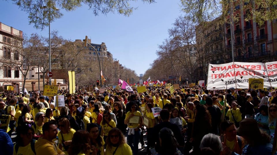 A large crowd of people marching through a city street, holding banners and flags, with historic buildings in the background,