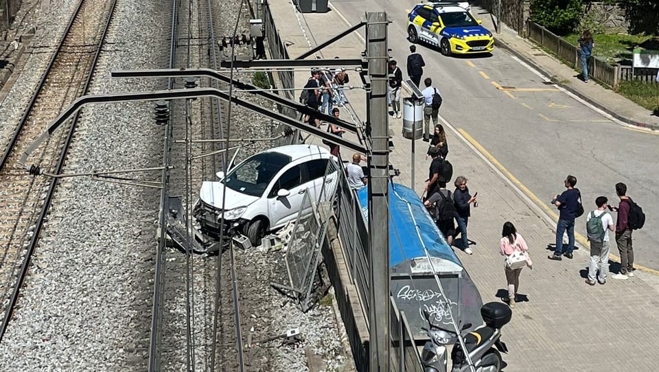 A damaged car on railway tracks next to a collapsed wall, with emergency services present.