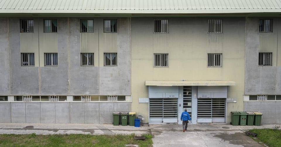 Prison guard standing in a corridor of a modern prison facility, looking towards the camera with a serious expression.