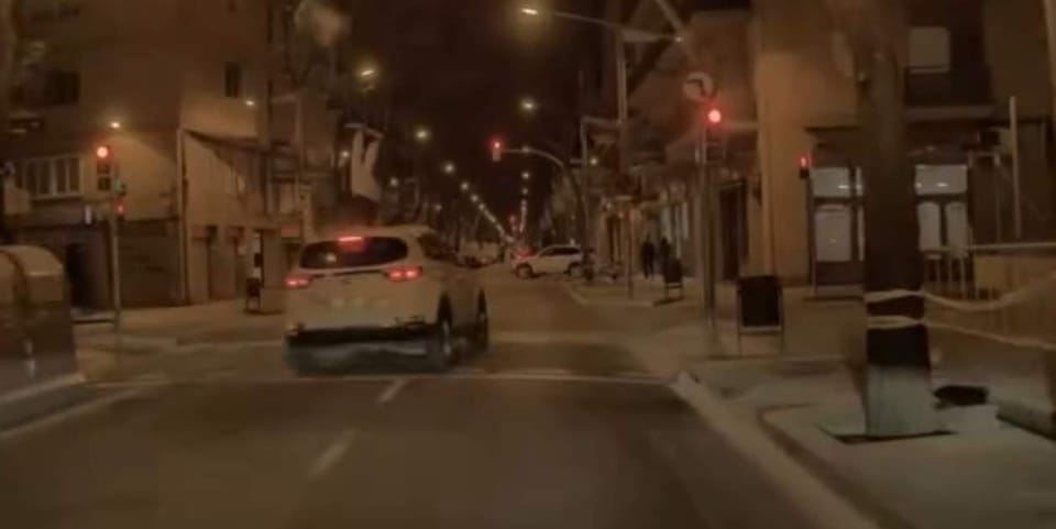 A white car colliding with a stationary motorbike at a red traffic light on a city street.