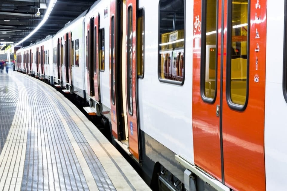 A modern FGC train arriving at a station platform, with passengers waiting. The train is white and red.