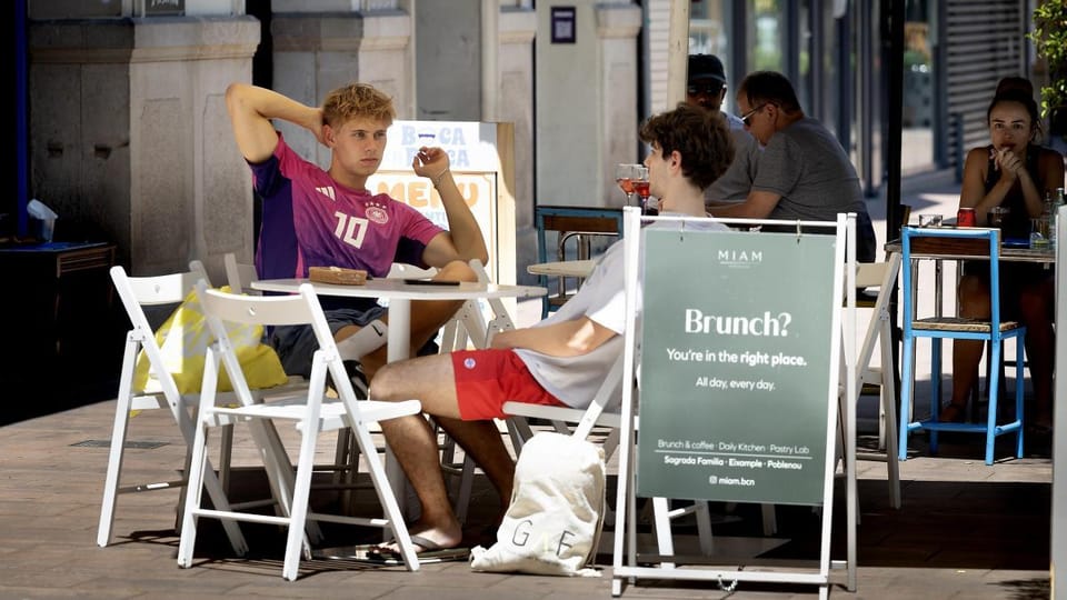 A modern cafe with outdoor seating on a tree-lined street in Poblenou, Barcelona, with people enjoying brunch.